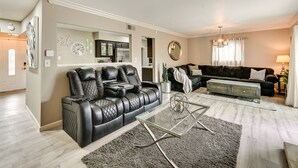 Ein modernes Wohnzimmer mit einem schwarzen Ledersofa, einem Glastisch und einem Essbereich, im Hintergrund ist eine Küche zu sehen.