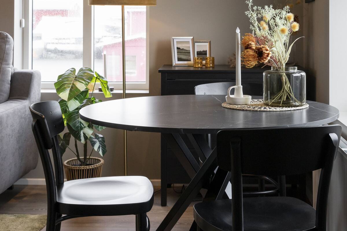 A dining area with a round table, black chairs, a potted plant, a tall floor lamp, and a vase with dried flowers.
