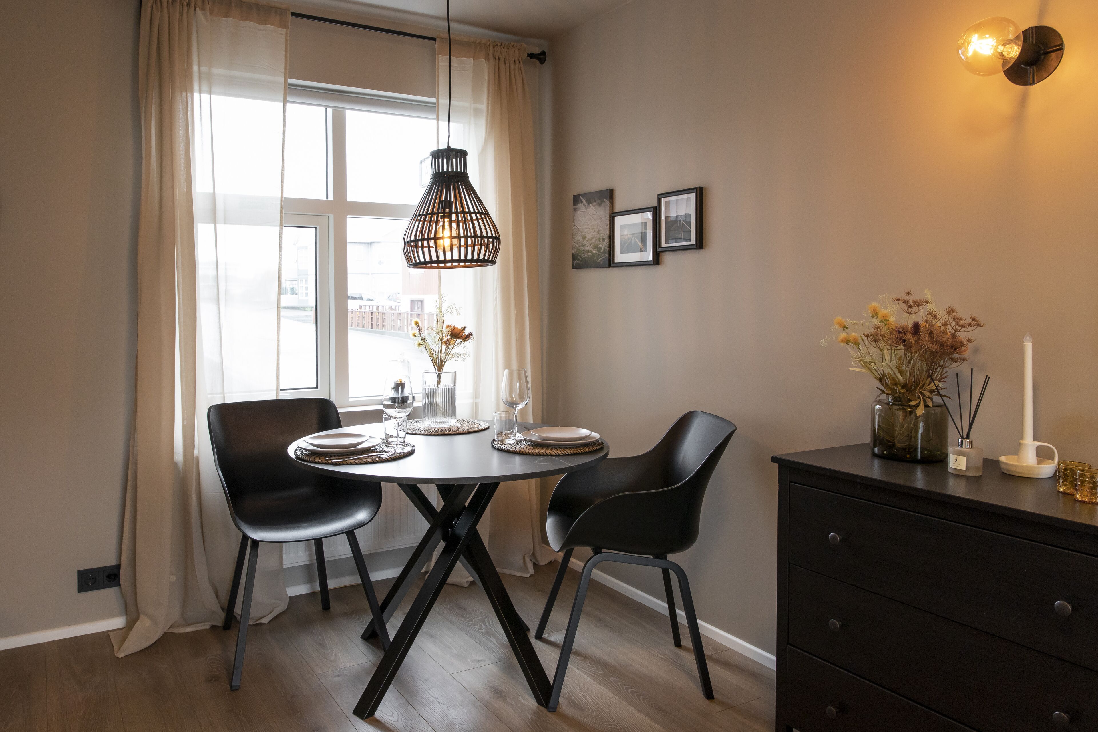 A dining area with a round table, two chairs, a hanging light fixture, a vase with flowers, and a wall-mounted picture frame.