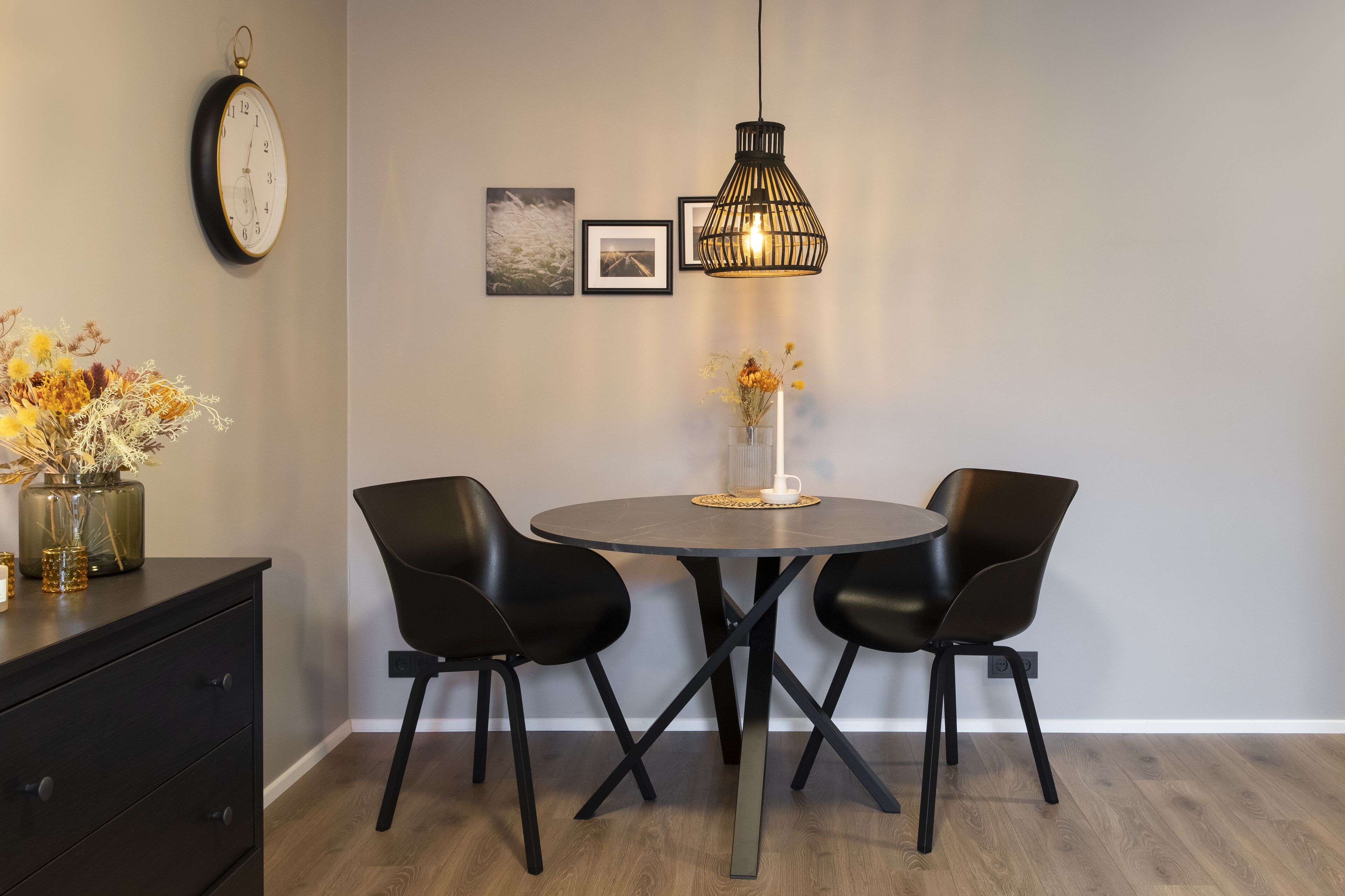 Zona de menjador amb taula rodona, dues cadires negres, llum penjant negra, rellotge de paret i quadres emmarcats.