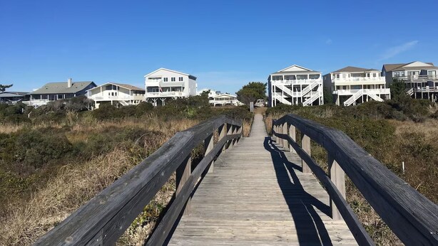 Ein Holzsteg führt zu Strandhäusern mit mehreren Stockwerken und Balkonen.