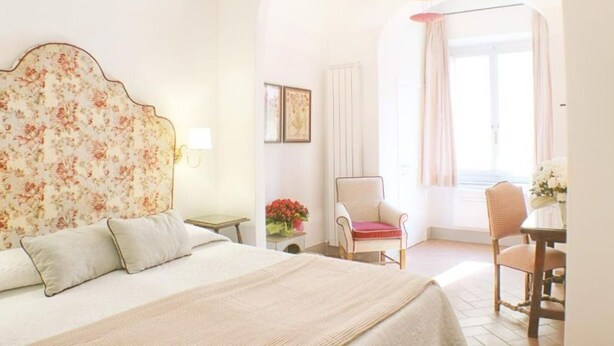 Ein Schlafzimmer mit einem blumigen Kopfteil, einem Bett mit Kissen, einem Nachttisch, einem Sessel, einem Tisch mit Blumen und einem Fenster mit Vorhängen.