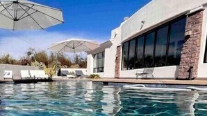 A clear swimming pool with lounge chairs and umbrellas, surrounded by a modern building with large windows and stone accents. - Pristine Luxury Estate, Views, Sleeps 26, Heated Pool & Spa (Fountain Hills)