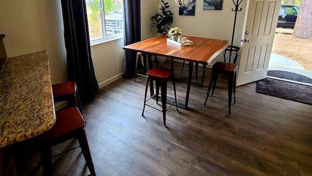 A dining area with a wooden table, four chairs, a chandelier, and a view of the outdoors through a window.