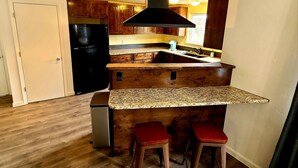 A compact kitchen with wooden cabinets, a black range hood, a granite countertop, and two red bar stools.