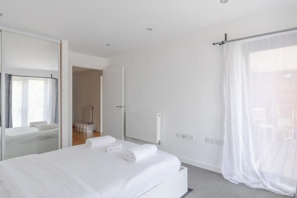 A modern hotel room with a large bed, a bathroom with a shower, and a view through a window with sheer curtains. - Vibrant 1BD Flat w/ Private Balcony, Camberwell! (London)
