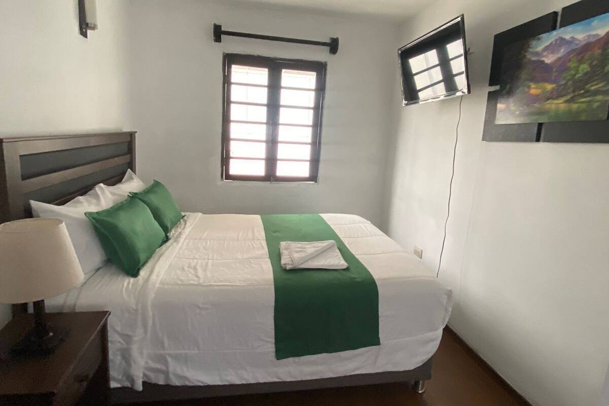 A neatly made bed with a green throw, a bedside lamp, and a window with blinds.