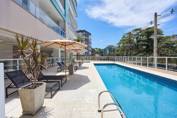 A swimming pool with lounge chairs and umbrellas, surrounded by a building with balconies. - Sunset (Ubatuba)