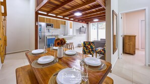 Un comedor con una mesa de madera dispuesta para cuatro, una cocina al fondo y una silla colorida.