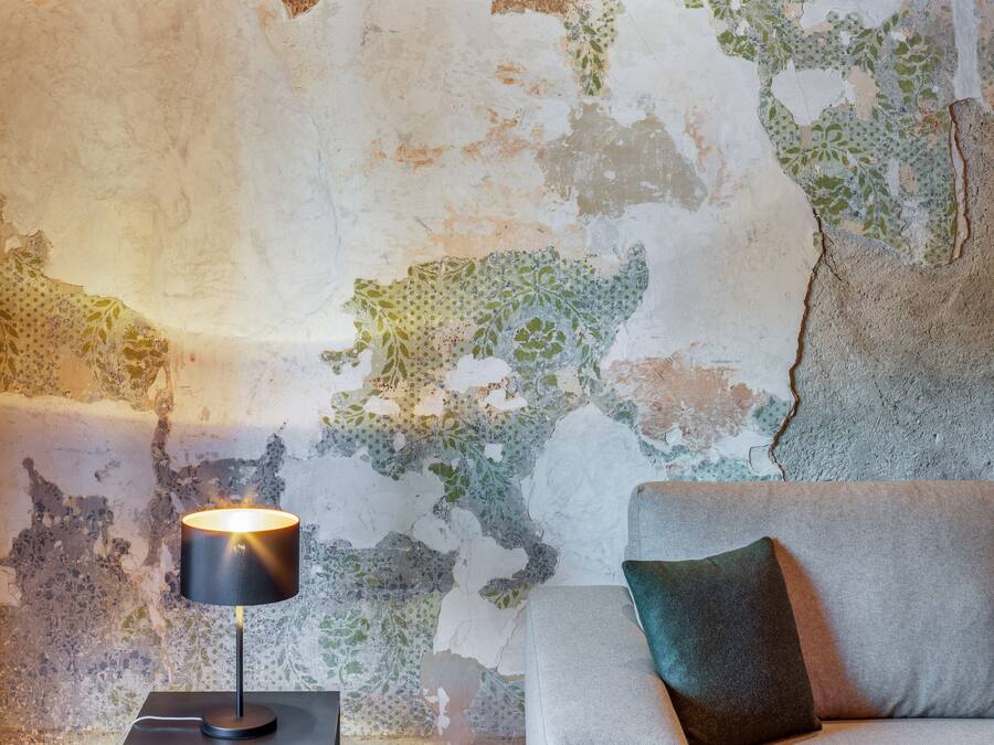 Una habitación con techo de madera, pared texturizada con pintura descascarada, una lámpara moderna sobre una mesita y un sofá gris.