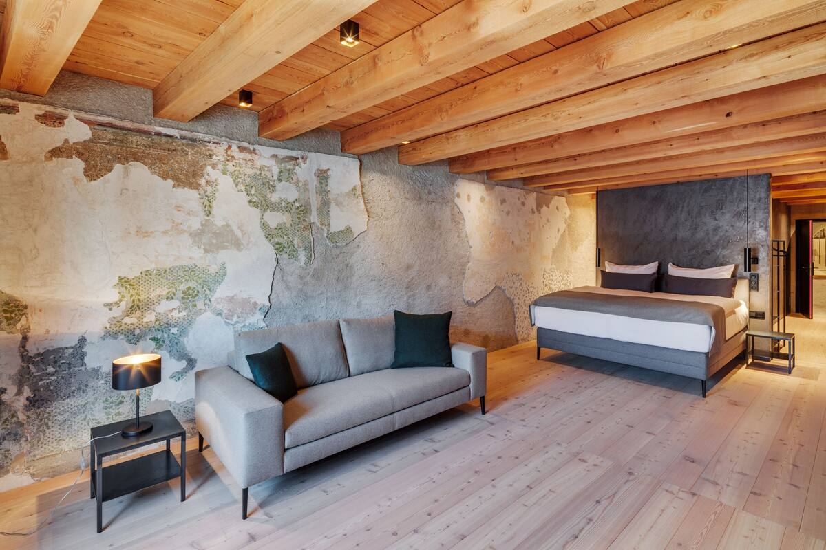 A bedroom with a wooden ceiling, a bed, a sofa, and a nightstand with a lamp.