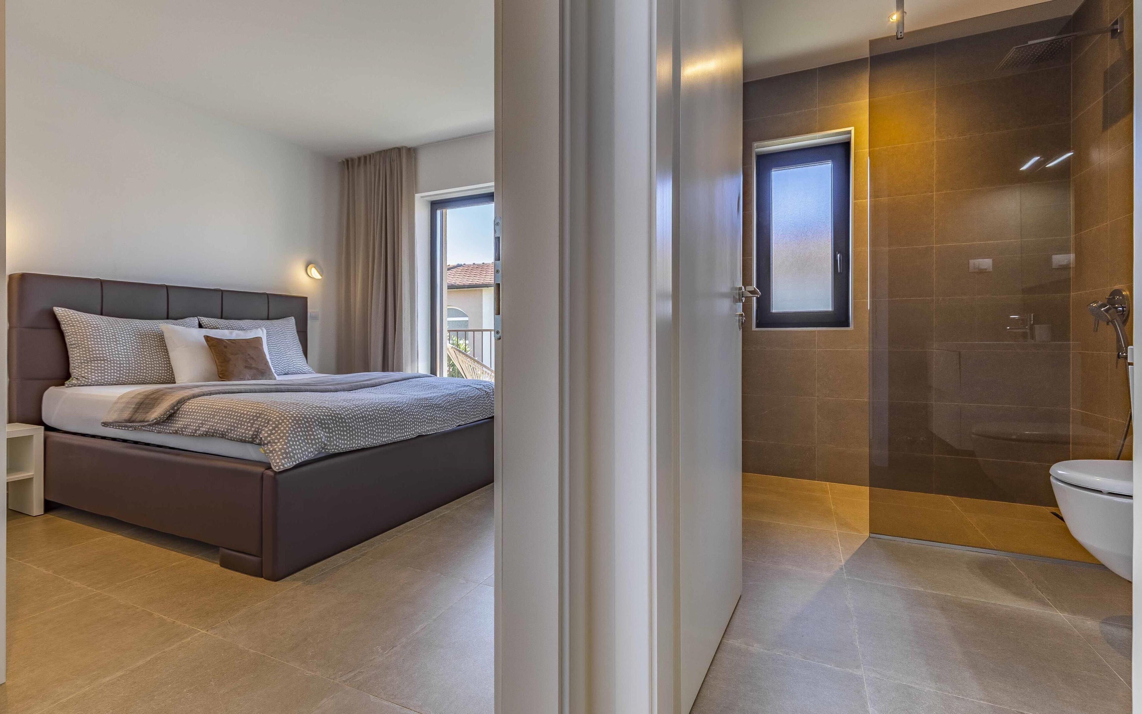 A modern bedroom with a large bed, a nightstand, and a view of the outside. Adjacent to the bedroom is a bathroom with a shower, toilet, and a glass-enclosed shower area.