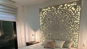 Une chambre à coucher avec un grand lit, une tête de lit décorative, une table de chevet, une lampe et une vue sur l’extérieur, accessible par une porte coulissante.