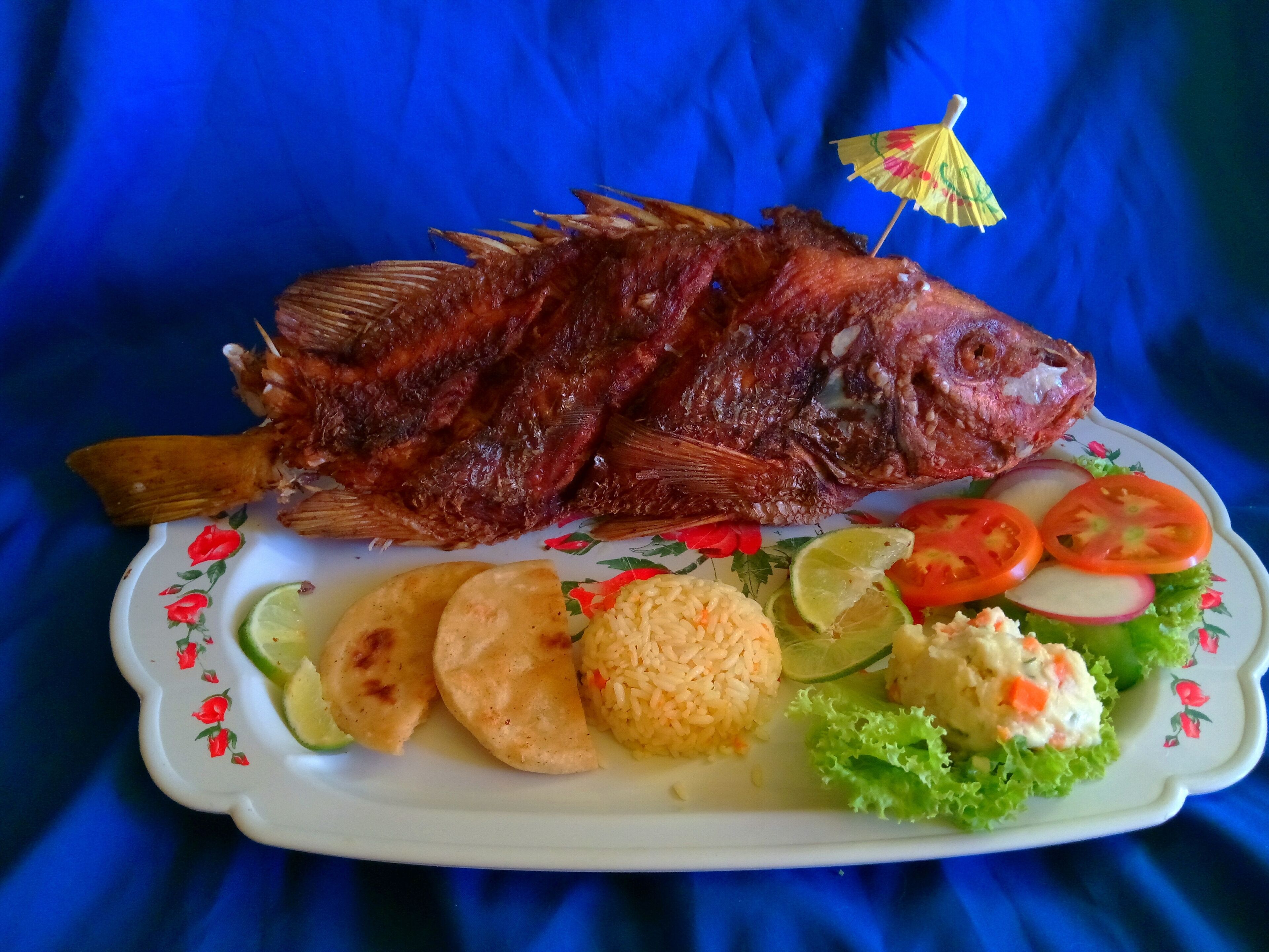 Un plato con un pescado frito entero, cuñas de lima, tomates en rodajas, lechuga y acompañamiento de arroz y patatas fritas.