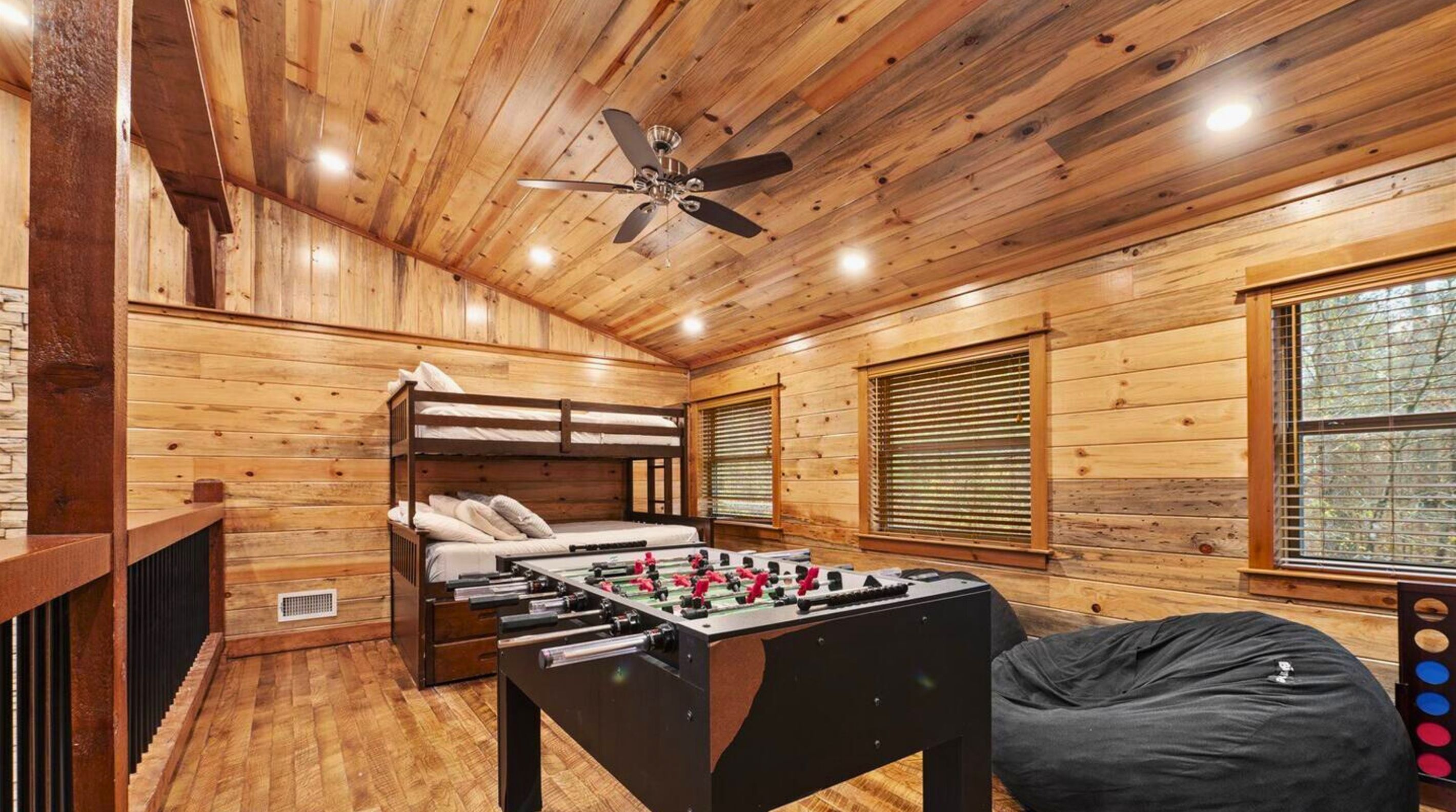 Une pièce aux murs en bois, avec un ventilateur de plafond, un lit, une table de baby-foot et un fauteuil poire.