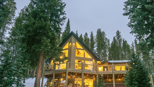 A large log cabin with multiple balconies and large windows, surrounded by tall pine trees and snow.