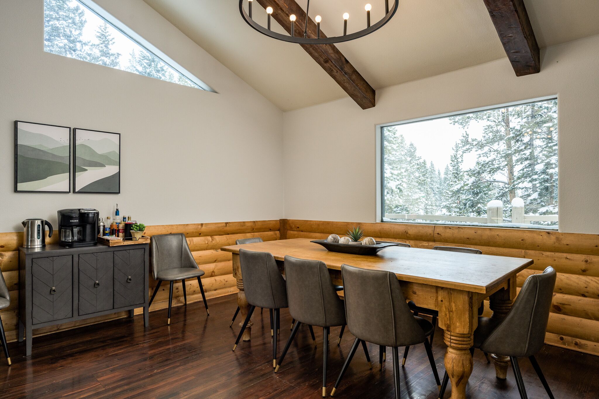A cozy dining area with a wooden table and chairs, a built-in bench along the wall, and a large window offering a view of snow-covered trees.