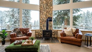 A cozy living room with a stone fireplace, leather sofas, and a large window overlooking a snowy forest.