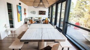 Een moderne eetruimte met een grote tafel, bankjes en stoelen. Er hangen kunstwerken aan de muren, een bank en een uitzicht door grote ramen.