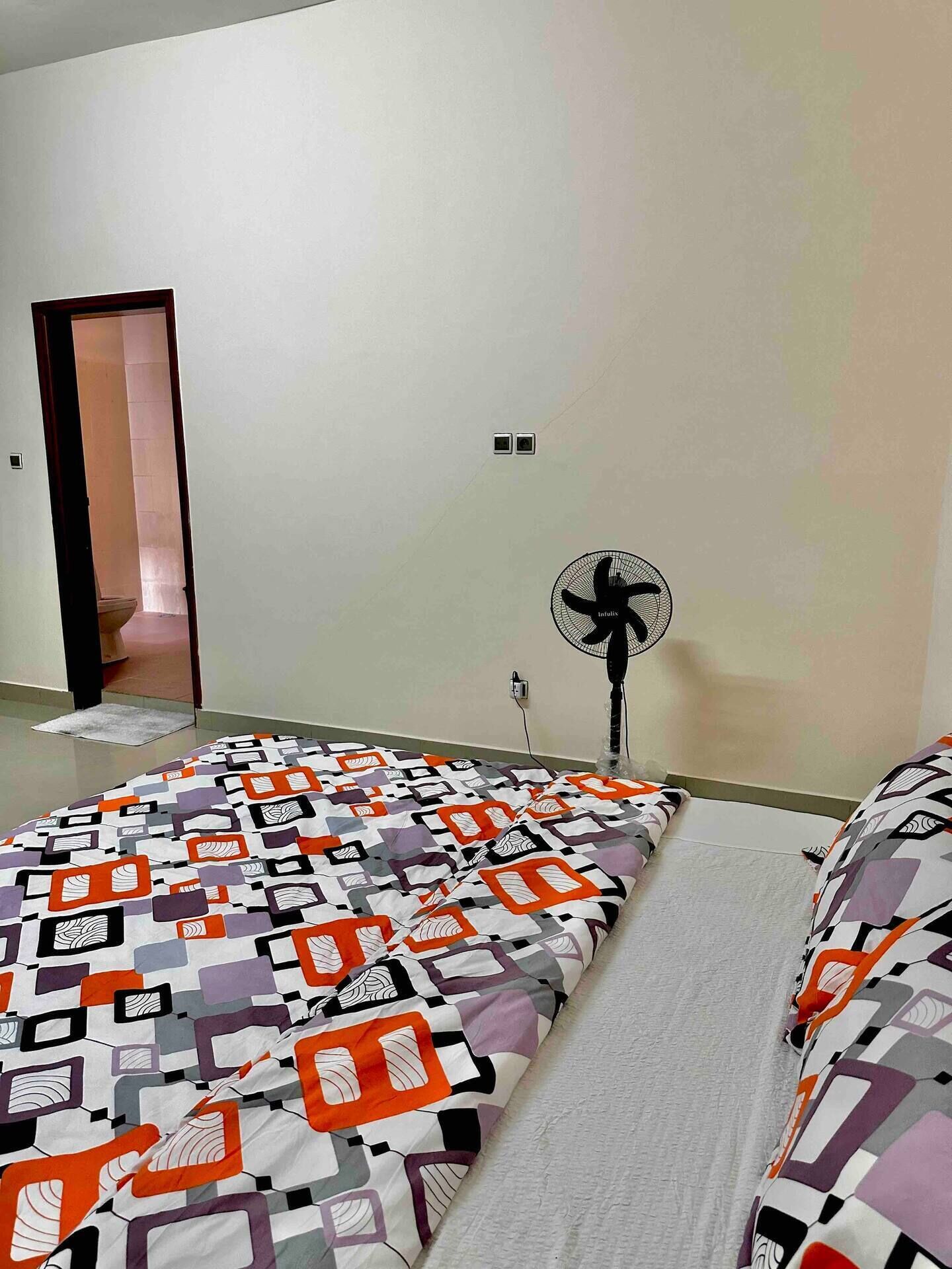 A bedroom with a patterned bedspread, a standing fan, and a door leading to another room.