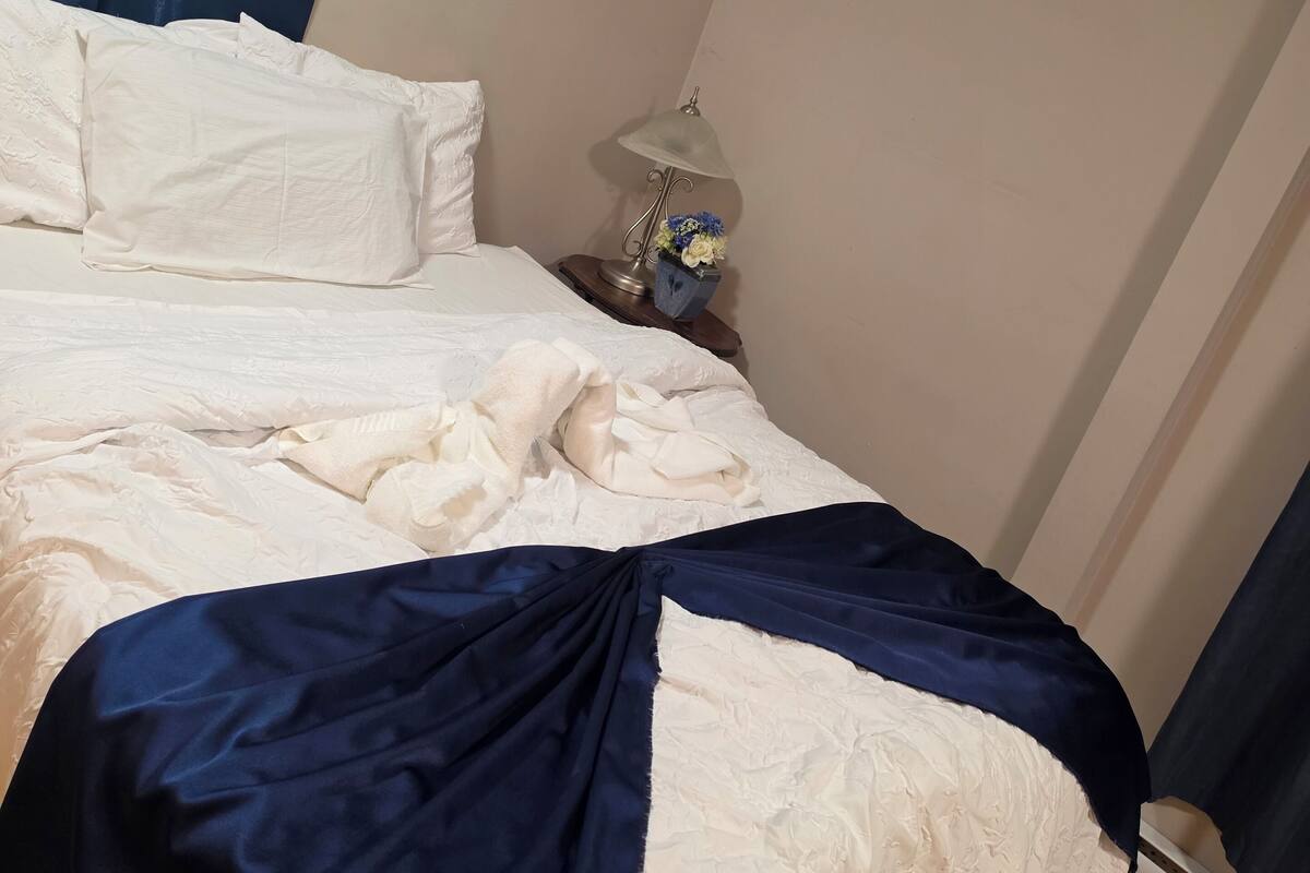 A neatly made bed with a white and blue bedspread, a bedside table with a lamp and flowers, and a wooden floor.