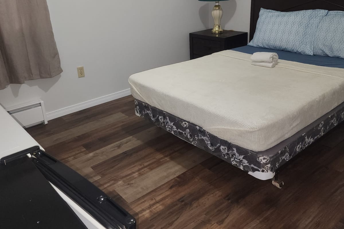 A bedroom with a bed, a nightstand with a lamp, a patterned bed skirt, and a window with a curtain.