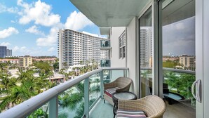 A balcony with wicker furniture and a glass railing overlooking a cityscape with palm trees and high-rise buildings. - Direct access to beach and heated pool at The Tides (Hollywood)