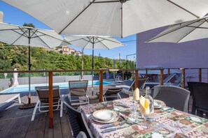 A poolside dining area with outdoor seating, umbrellas, and a view of a pool and hillside buildings.