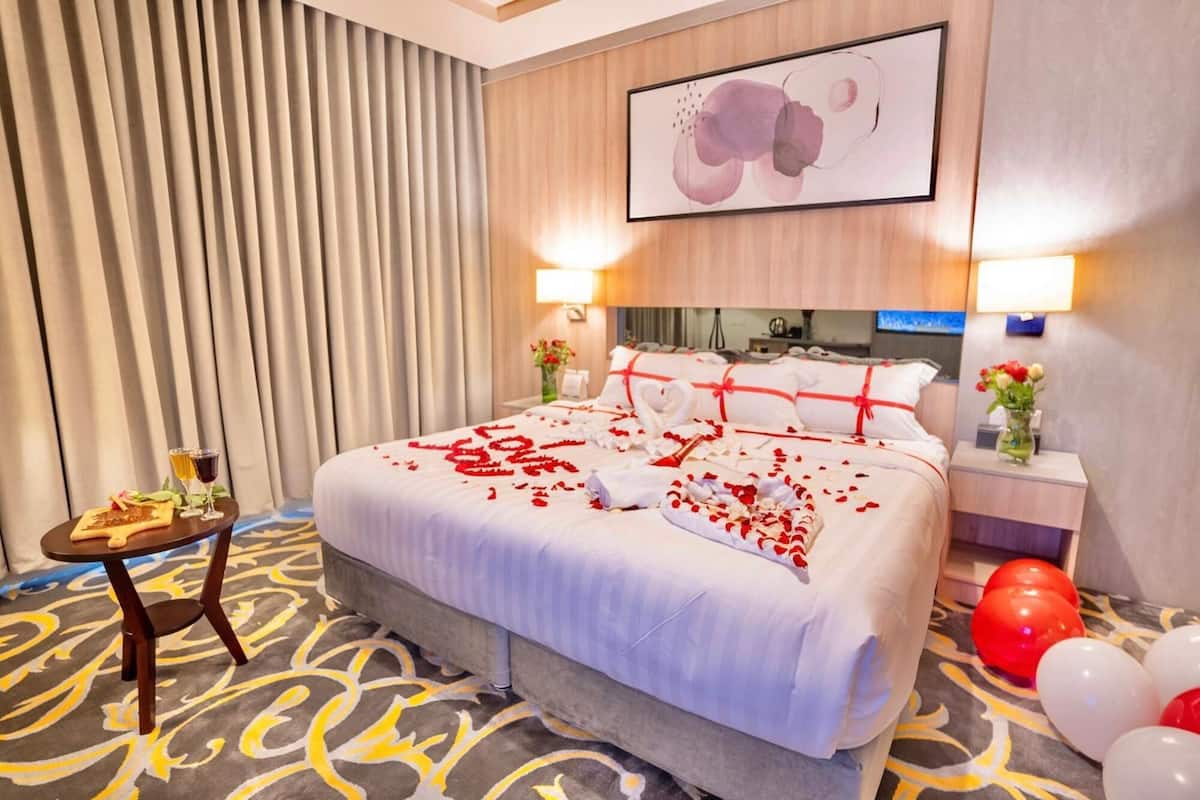 A hotel room with a bed adorned with rose petals and a heart-shaped towel, a small table with drinks and snacks, and a vase with flowers.