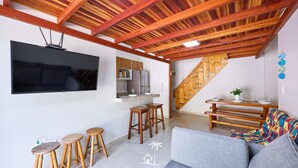A modern living space with a wooden ceiling, a flat-screen TV mounted on the wall, a kitchen area with a refrigerator, and a staircase leading to another level. - Brisamar (Ubatuba)