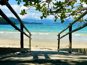 Une terrasse en bois qui mène à une plage bordée d’une eau turquoise et limpide, avec des montagnes qui se dessinent à l’horizon.