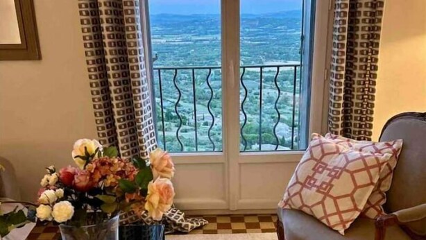 A room with a view of the sea, a chair with a patterned pillow, a vase with flowers, and a book on a table. - Gordes Idyllique Avec vue Panoramique (Gordes)