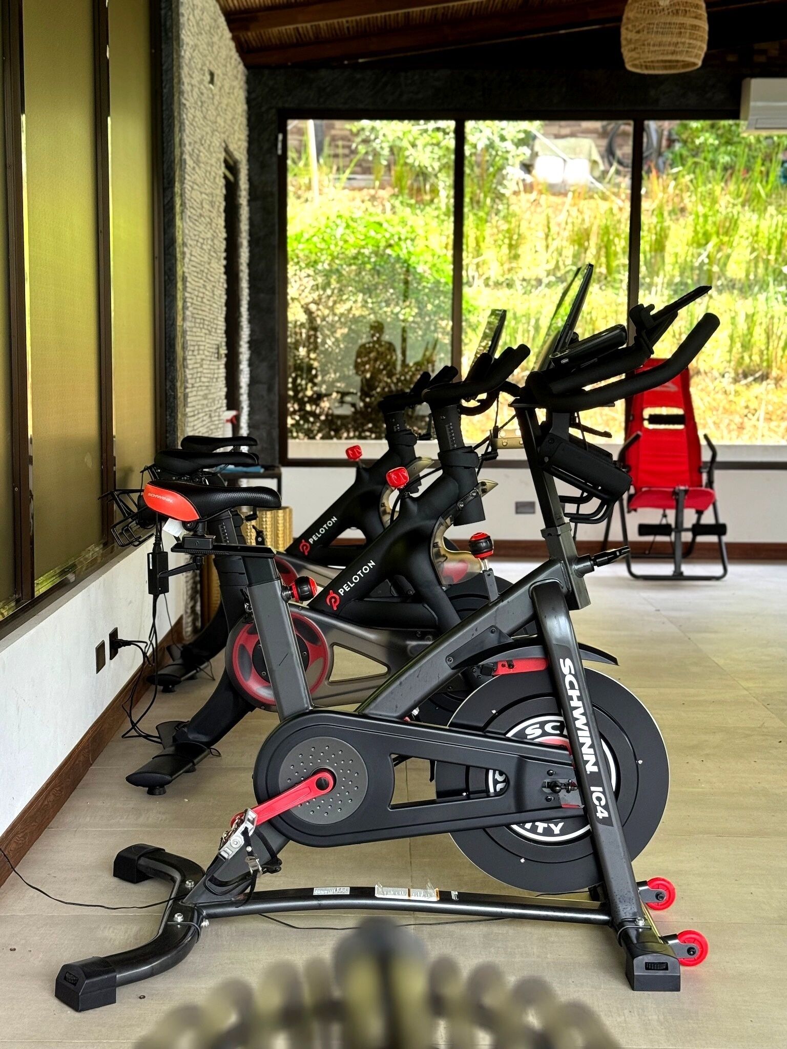 Una bicicleta estática con pantalla digital, una silla roja y una ventana con vista a follaje.