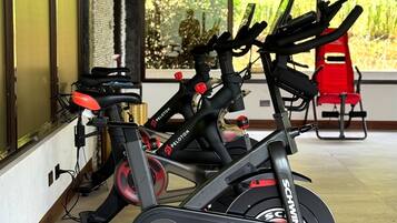 Una bicicleta estática con pantalla digital, una silla roja y una ventana con vista a follaje.