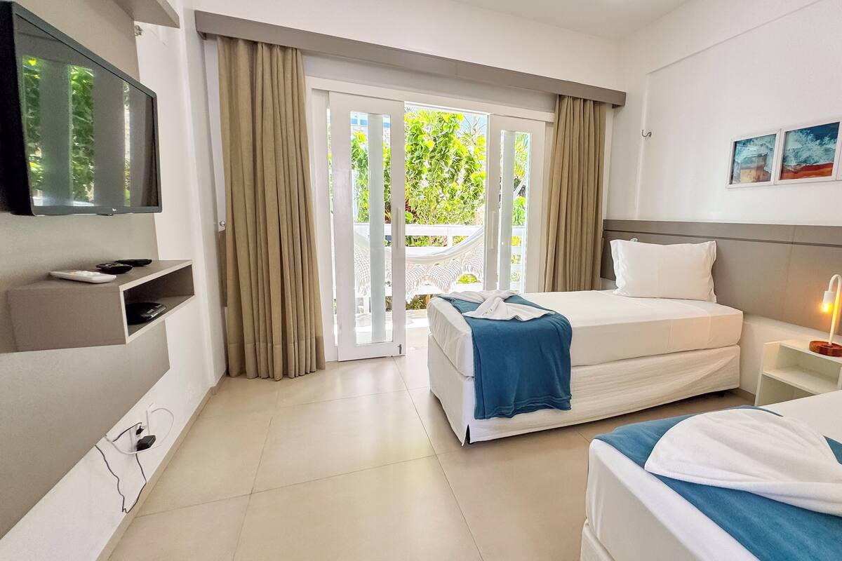 A hotel room with two beds, a TV, a bedside table with a lamp, and a view of greenery through the window.