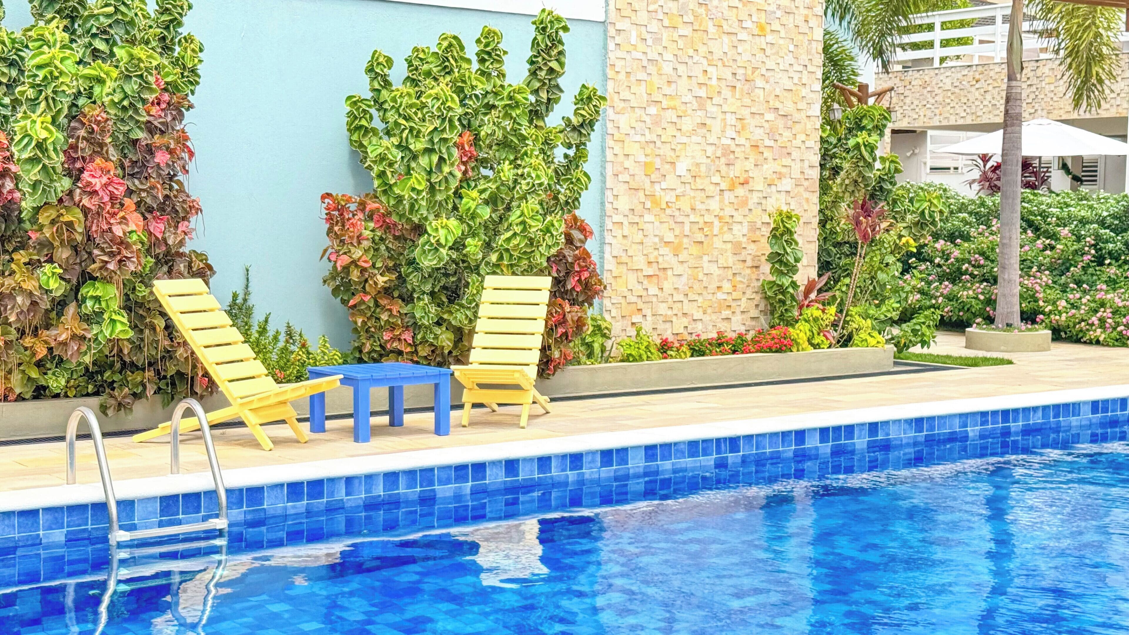 A swimming pool with two yellow lounge chairs and a small table, surrounded by lush greenery and a tiled wall.