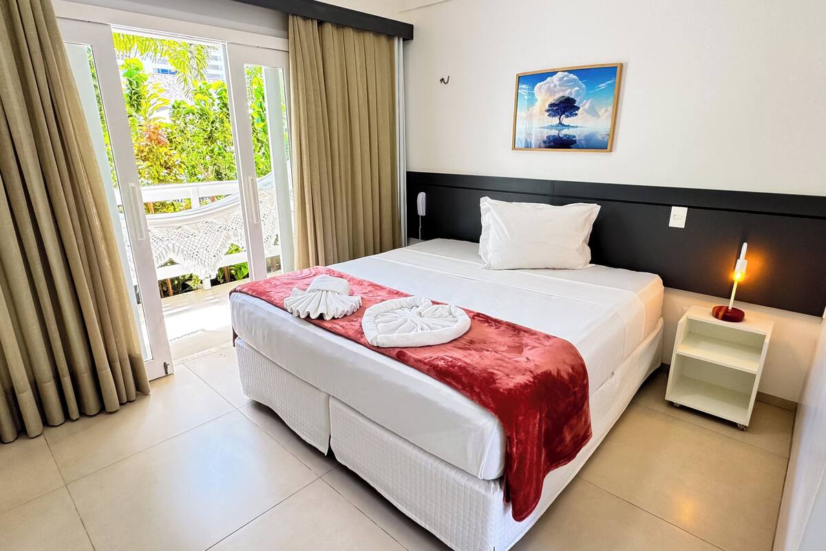 A hotel room with a bed, a nightstand, a painting, and a view of greenery outside.