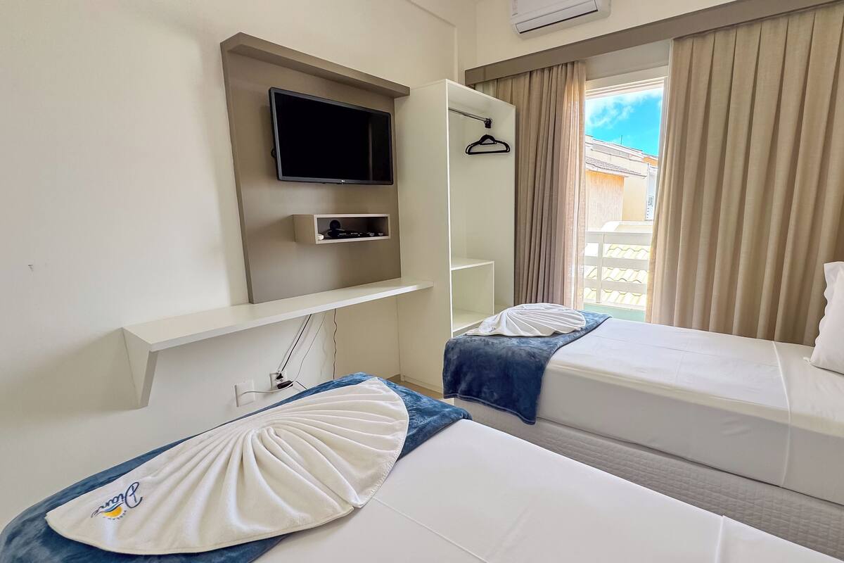 A hotel room with two beds, a TV mounted on the wall, a window with curtains, and a view of the outside.