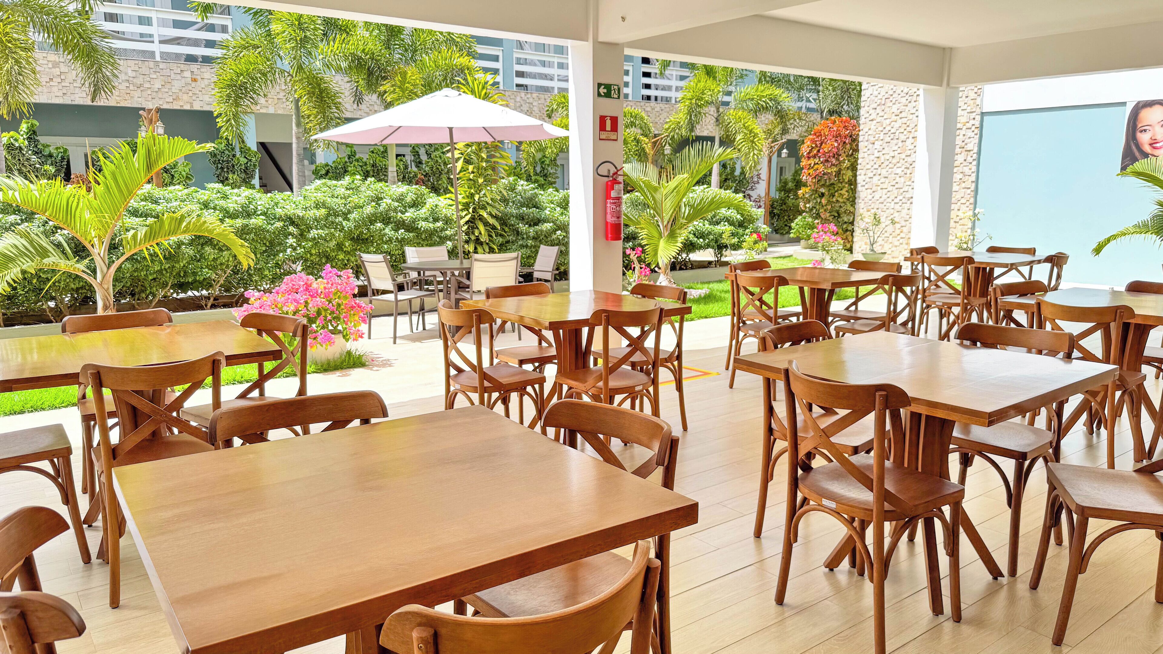 An outdoor dining area with wooden tables and chairs, surrounded by lush greenery and a view of a building in the background.