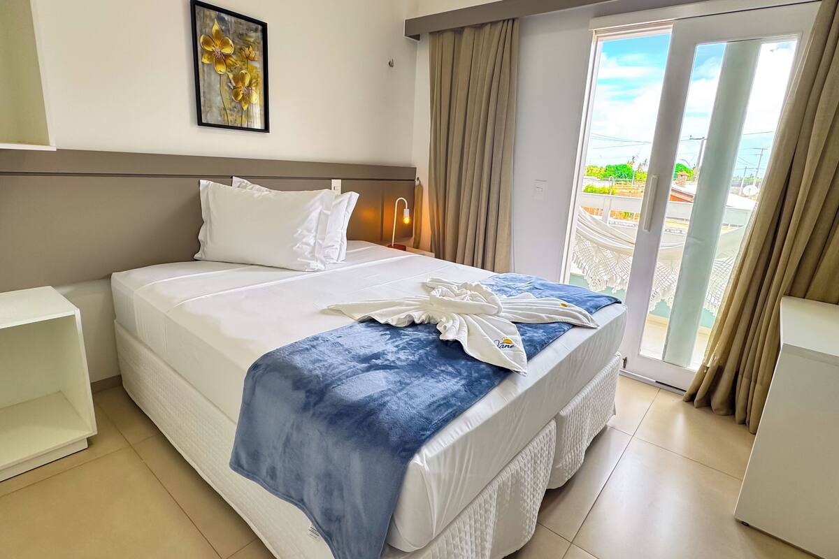 A hotel room with a bed, a nightstand, a window with curtains, and a view of the outside.