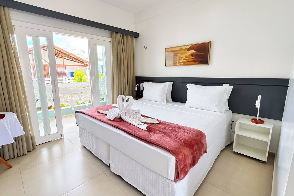 A hotel room with a bed, a nightstand, a table with a flower, and a view of the outside.