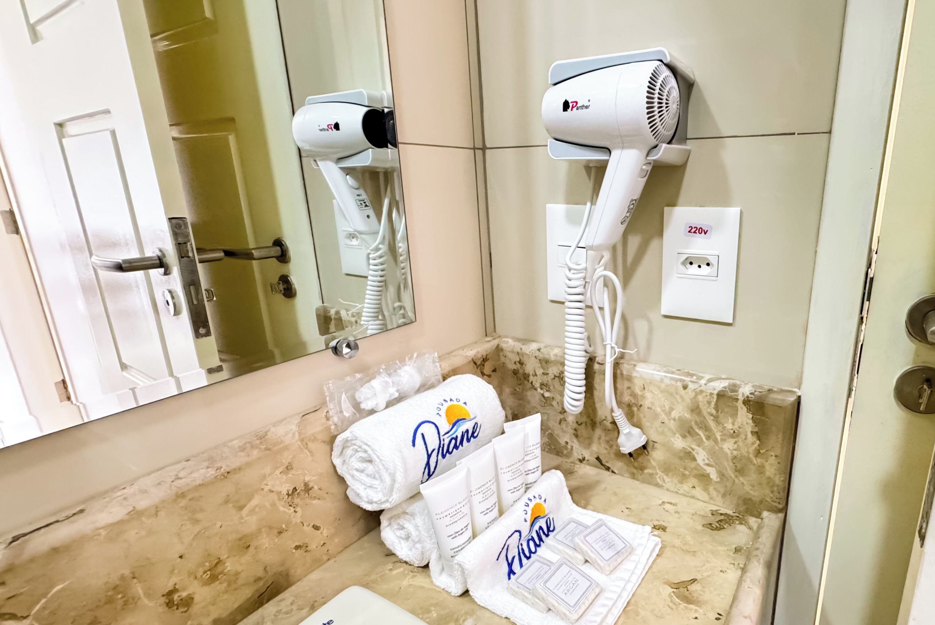 A bathroom with two hair dryers mounted on the wall, a towel rack with towels, and a 220V outlet.