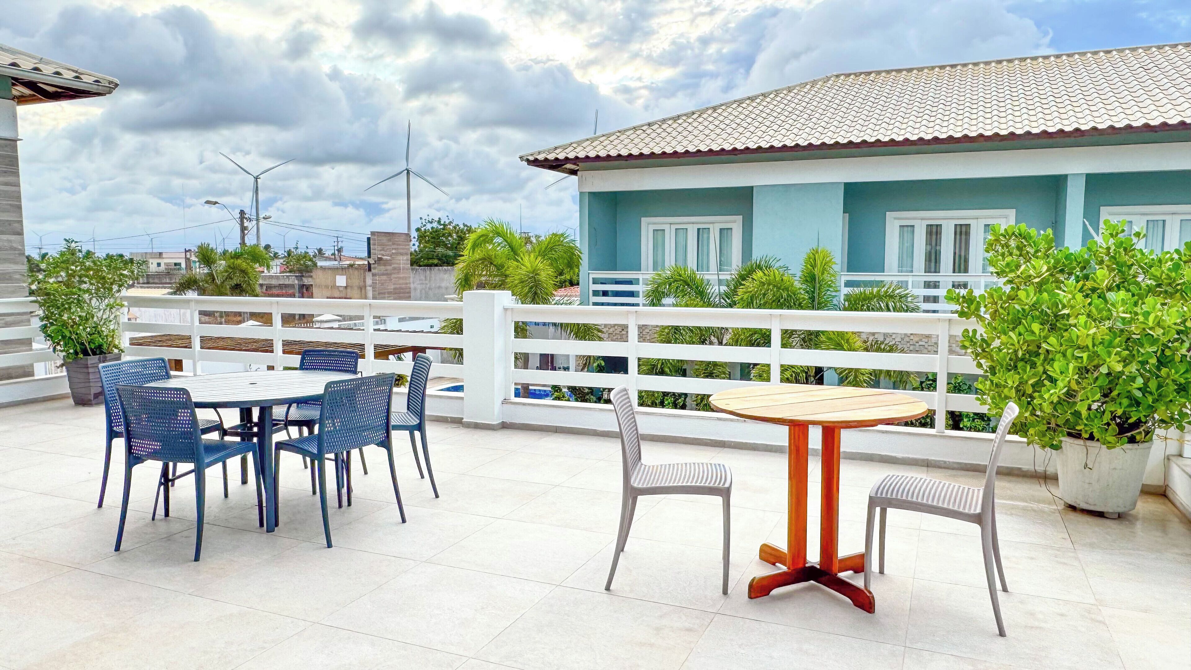 Una terraza en la azotea con una mesa y sillas, barandal blanco y un edificio con tejado de baldosas al fondo.