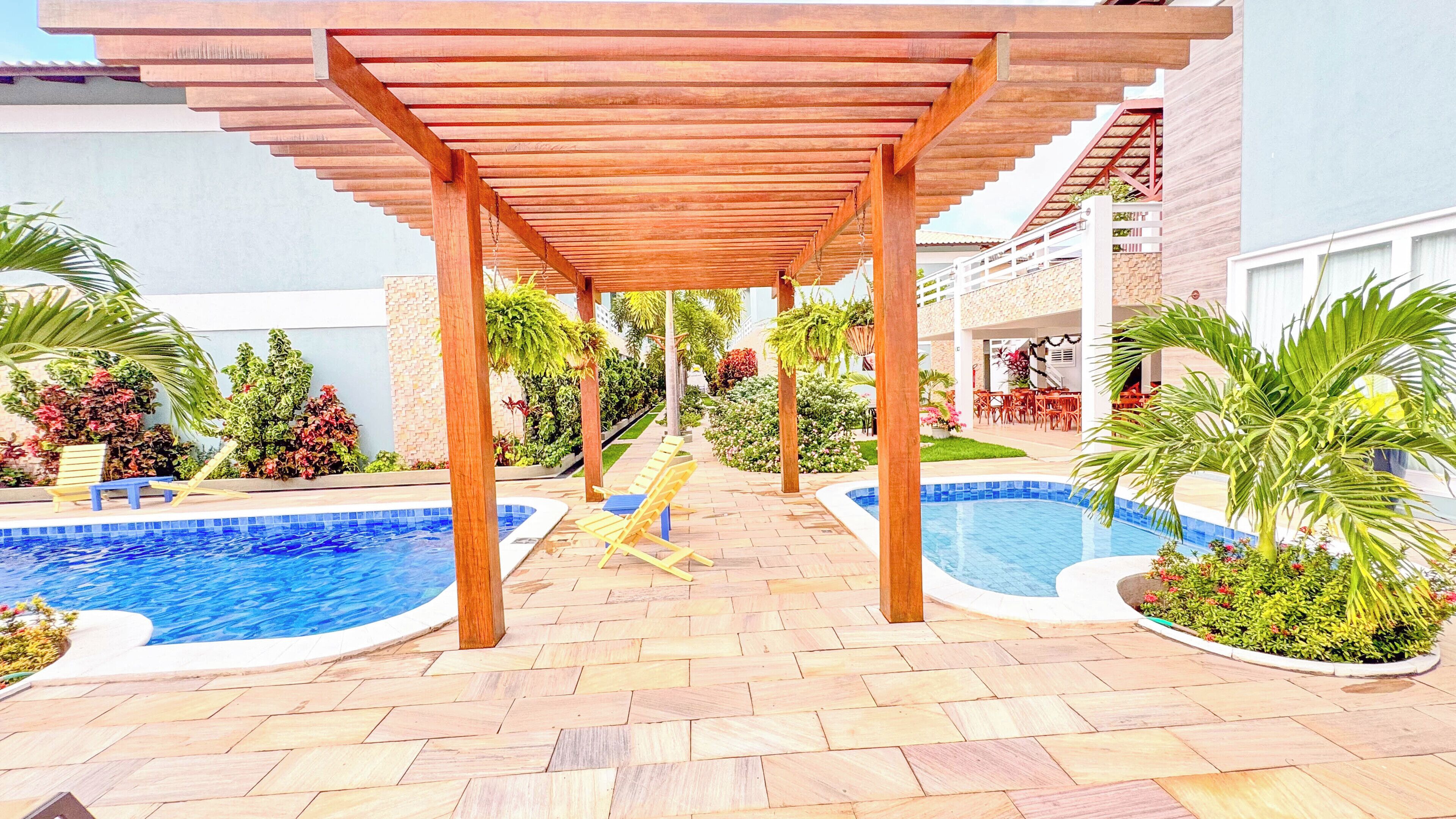 A covered outdoor area with two swimming pools, wooden pergolas, and various plants.