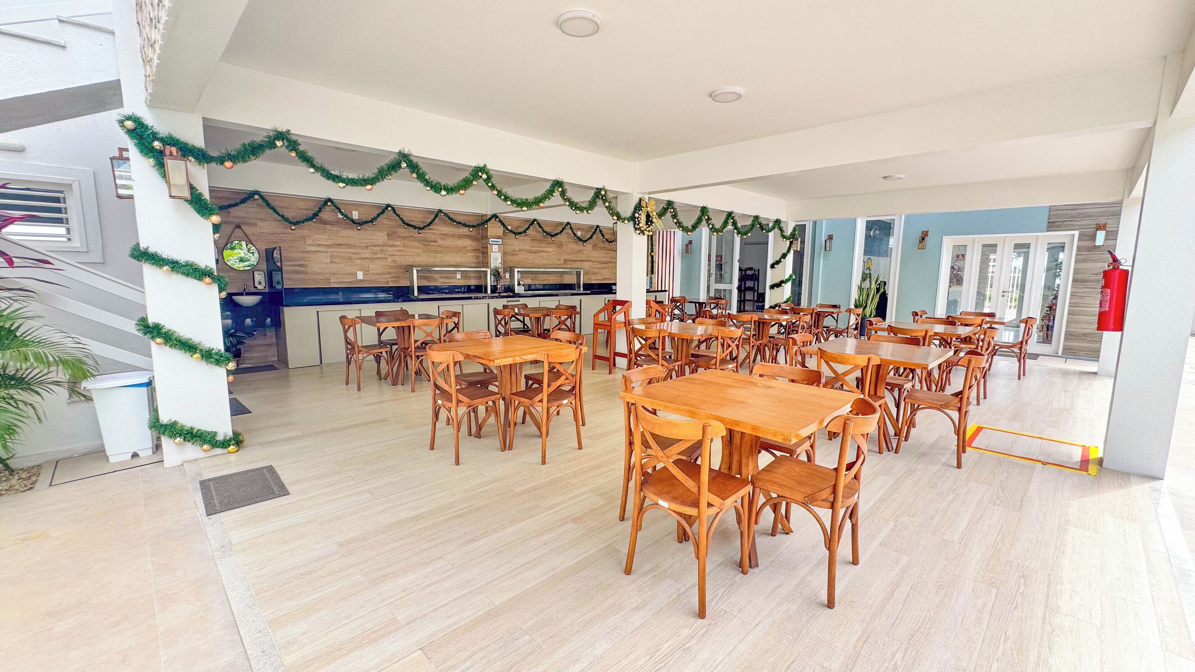 Un grand espace dédié à la salle à manger, avec des tables et des chaises en bois, une cuisine à l’arrière-plan et des guirlandes décoratives accrochées aux murs.
