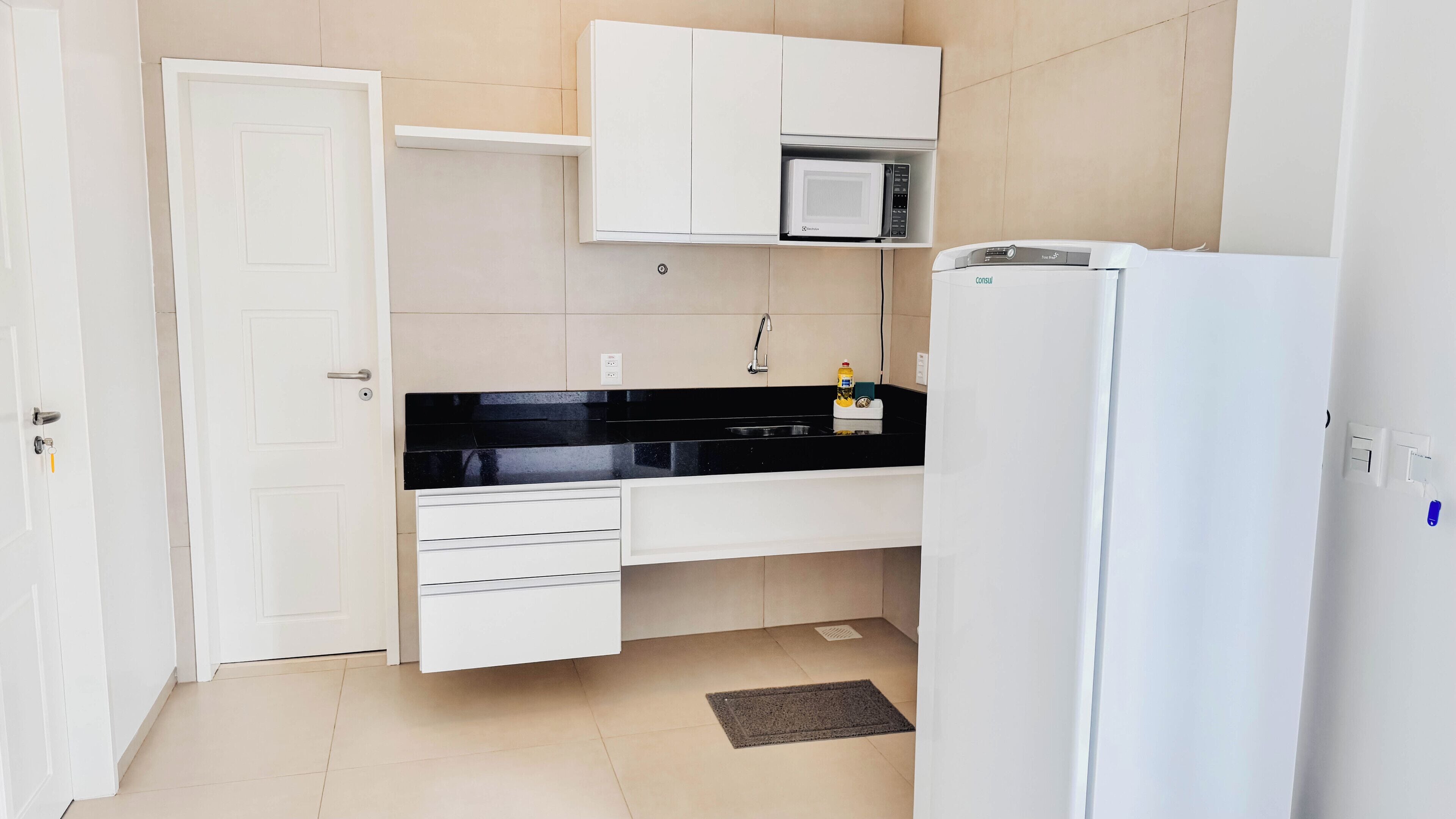 Una cocina compacta con muebles blancos, una encimera negra, microondas, fregadero y refrigerador.