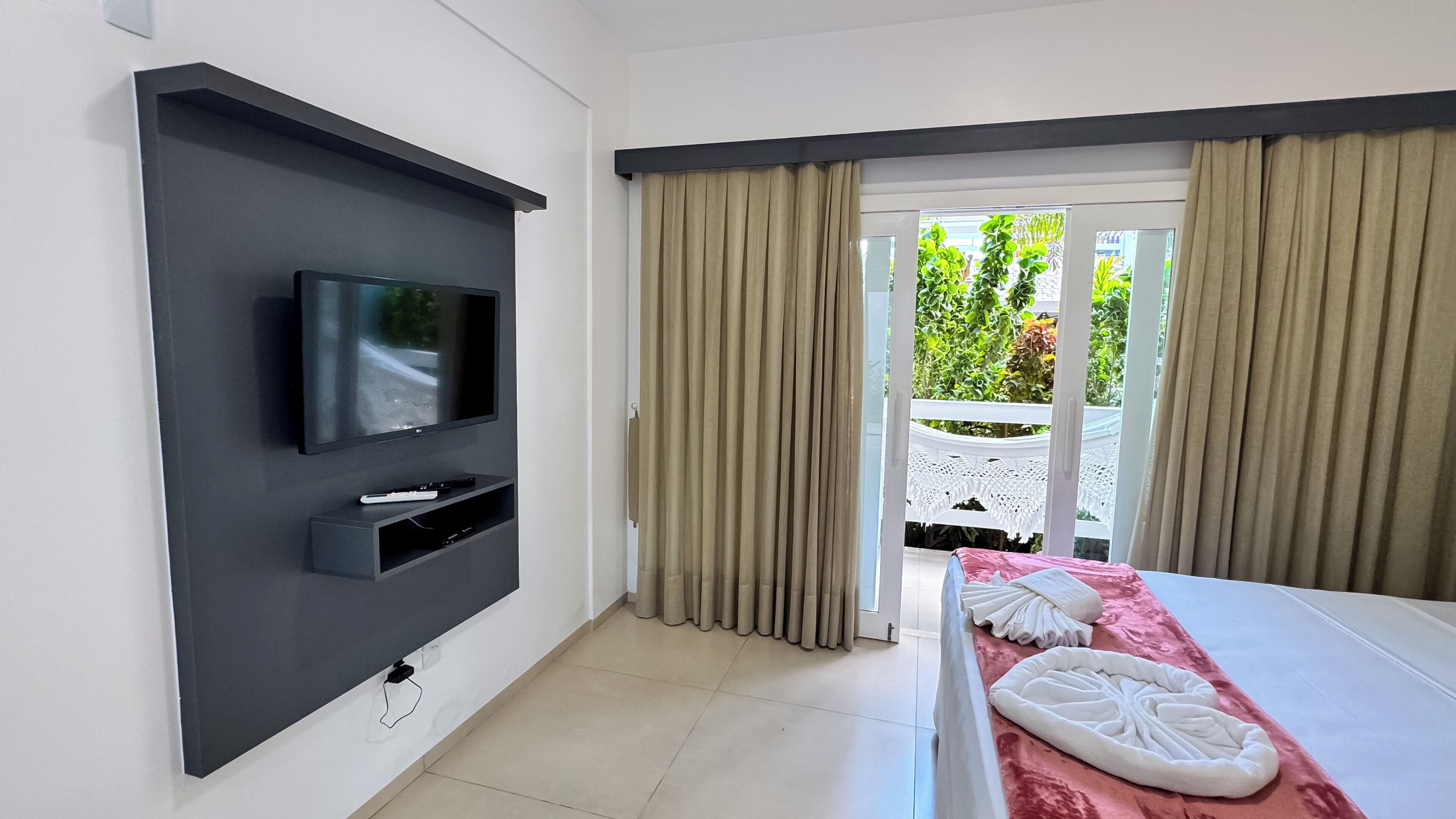 A modern hotel room with a flat-screen TV mounted on the wall, a bed with a pink bedspread, and a balcony with beige curtains.