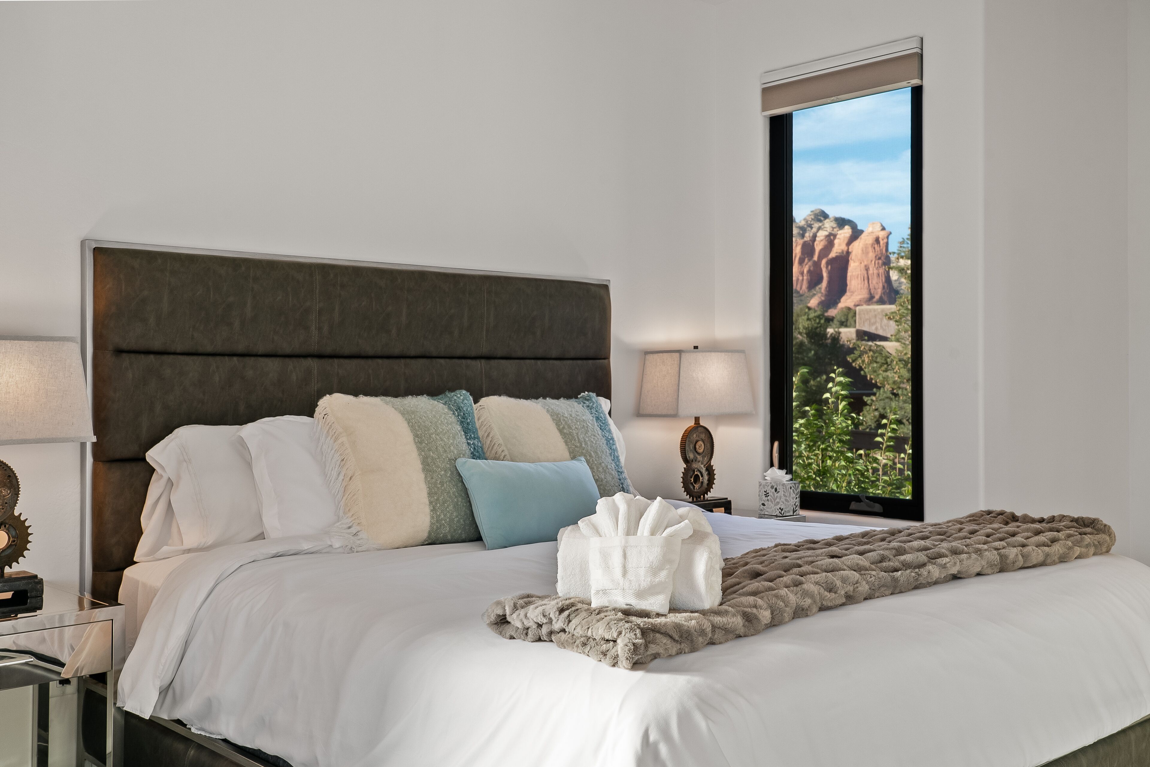 Un lit bien fait, avec une couette blanche et un plaid marron, une tête de lit sombre et une vue sur des formations rocheuses rouges, visible par la fenêtre.