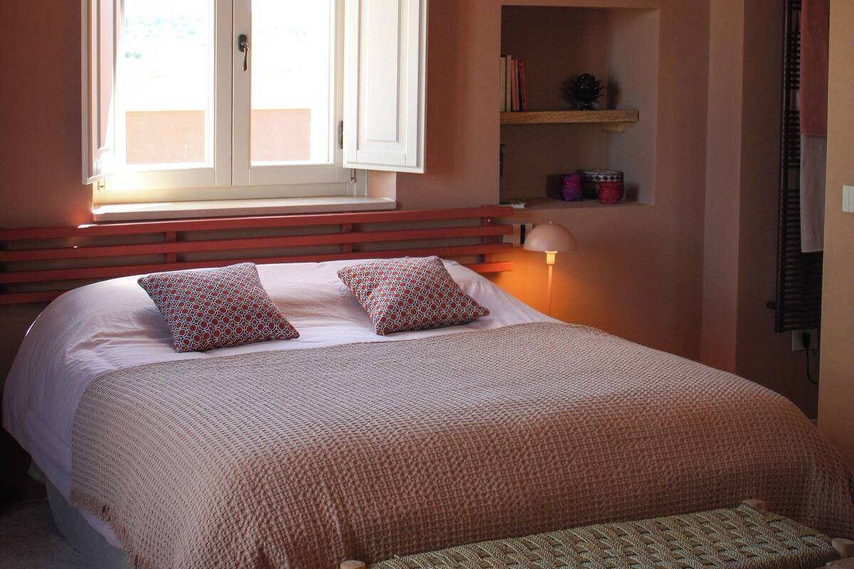 Een slaapkamer met een groot bed, een nachtkastje met een lamp, een raam met open luiken, en een plank met boeken en decoratieve objecten.