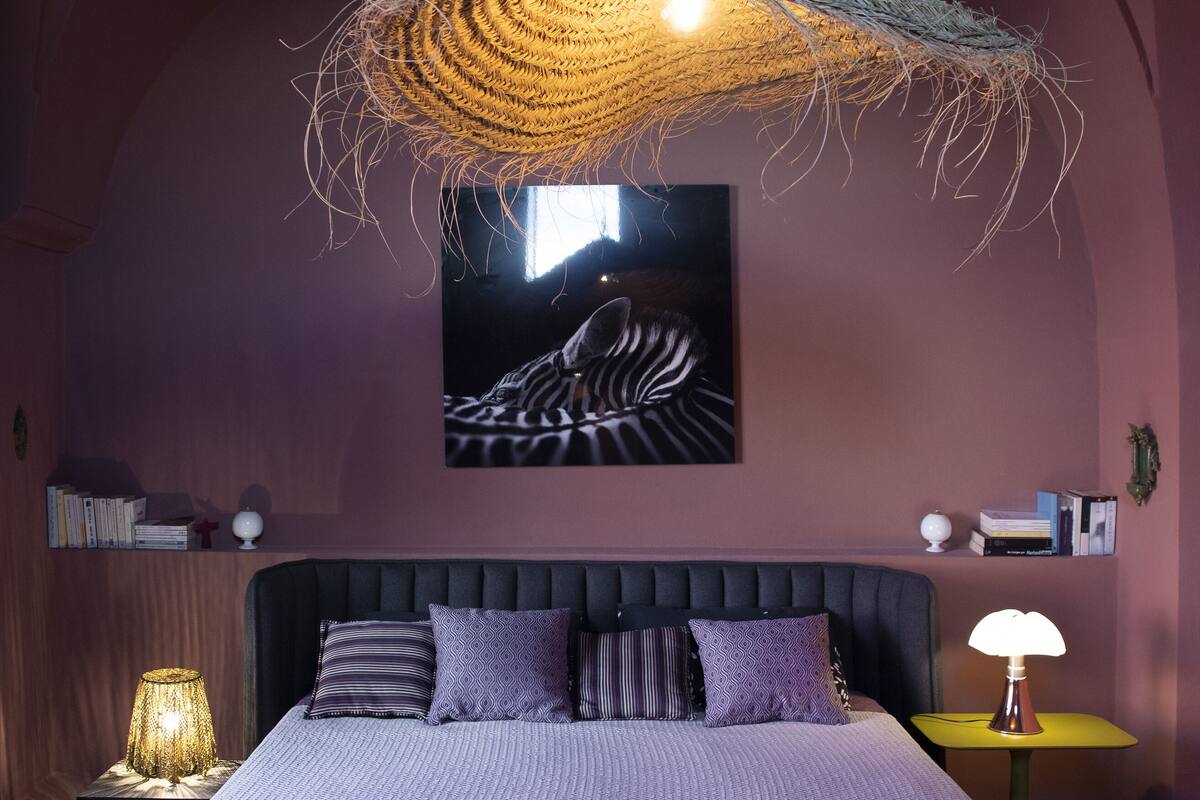 Een slaapkamer met een groot bed, een hanglamp, een aan de muur bevestigd schilderij en een nachtkastje met een lamp.